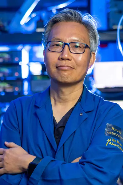 A man in a blue lab coat and glasses stands in a blue-lit laboratory. He has gray hair and a neutral expression, looking forward.